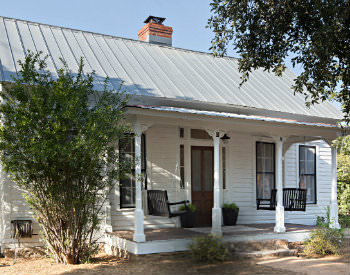 White cottage with metal roof and chimney, covered front porch with two porch swings amidst blue skies