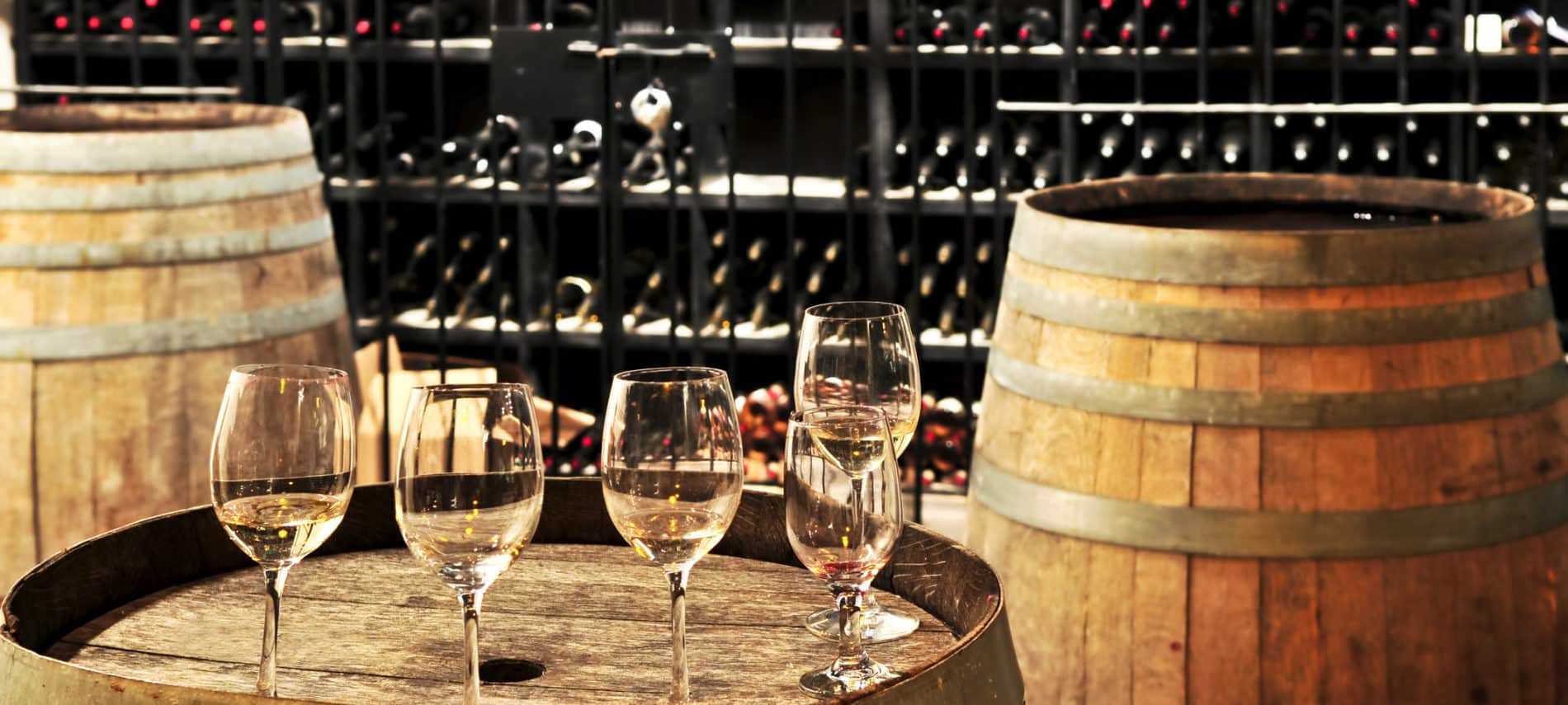 5 wine glasses with white wine on top of wine barrel, 1 wine barrel on each side, wine racks behind full of wine bottles